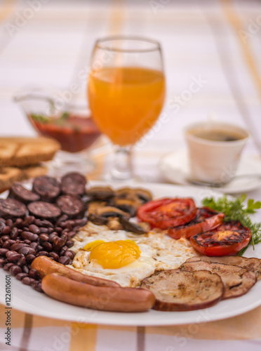 Full English fried breakfast with bacon, egg, sausages, black pudding, mushrooms, grilled tomatoes and baked beans. Closeup with toasts, cup of coffee, orange juice and home made red sauce.