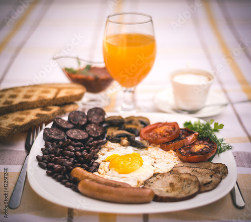 Full English fried breakfast with bacon, egg, sausages, black pudding, mushrooms, grilled tomatoes and baked beans. Closeup with toasts, cappuccino, orange juice and home made red sauce.