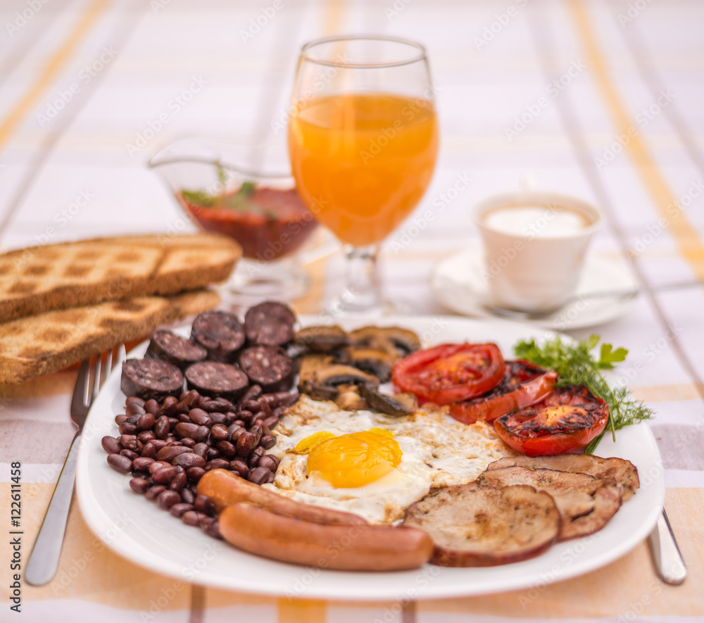 Full English fried breakfast with bacon, egg, sausages, black pudding, mushrooms, grilled tomatoes and baked beans. Closeup with toasts, cappuccino, orange juice and home made  red sauce.