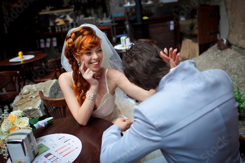 Funny red-haired bride smiles to her groom while he kisses her p