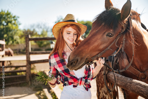 Cheerful woman cowgirl giving food to horse on ranch © Drobot Dean