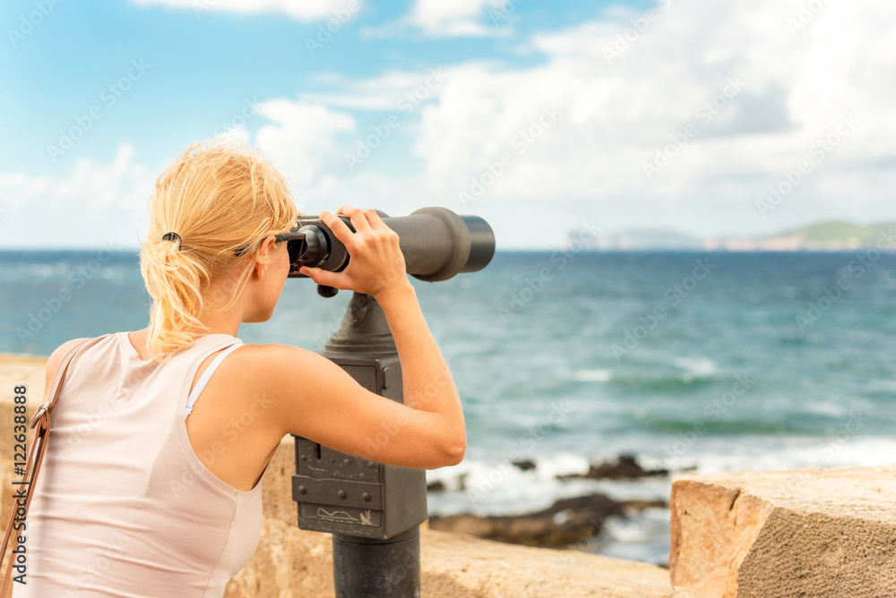 Young beautiful blonde woman using telescope to watch marine life and coast in Alghero, Sardegna, Italy