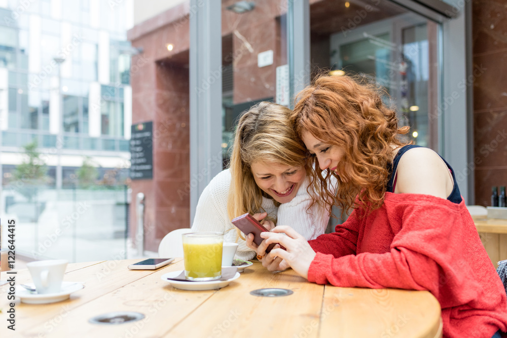 Two young beautiful caucasian blonde and redhead women using smart phone sitting in a bar - technology, social network, communciation concept Two young beautiful caucasian blonde and redhead women using smart phone sitting in a bar - technology, social network, communciation concept