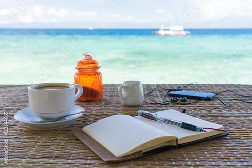Opened empty notepad is on the table with cup of coffee, phone and headphones at the tropical sea background