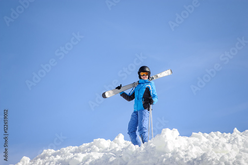Skier standing on top of the mountain against blue sky on a sunny day. Girl is holding skis on her shoulder smiling and looking into the camera. Winter sports concept. Carpathian Mountains