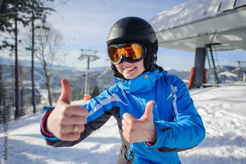 Close-up portrait of young happy female skier at ski resort smiling and showing thumbs up. Winter sports concept. Woman is wearing blue jacket, helmet and orange goggles. Bukovel, Ukraine