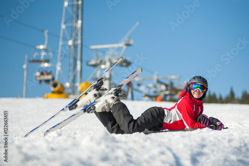 Woman smiling, lying with skis on snowy at mountain top in sunny day with ski lifts and blue sky in background.