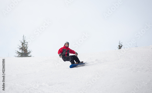 Young man snowboarder riding over the slope at the mountains overlooking a ski run on the snowy slope below at a winter resort, extreme sport