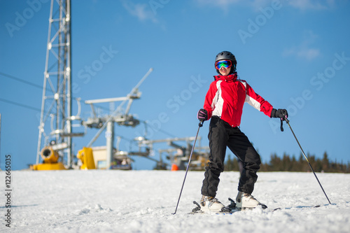 Woman wearing helmet, red jacket and ski goggles standing with skis on mountain top at a winter resort in sunny day with ski lifts and blue sky in background.
