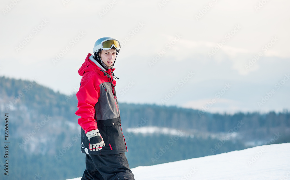 Portrait of snowboarder wearing helmet, red jacket, gloves and pants standing on top of a mountain and looking at the camera on the background of hills and the sky. Carpathian mountains, Bukovel