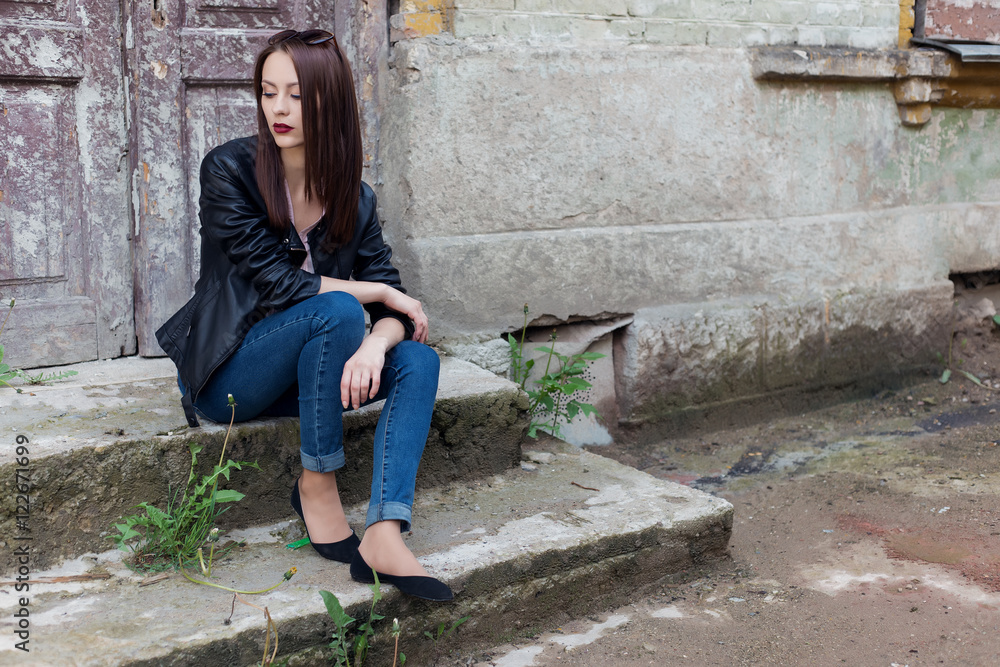 beautiful cute fashion girl with dark hair with sunglasses in a leather black jacket sitting on the stairs to the porch of the old buildings in the city beautiful cute fashion girl with dark hair with sunglasses in a leather black jacket sitting on the stairs to the porch of the old buildings in the city