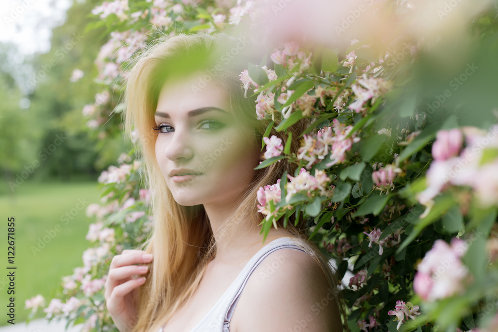 Fototapeta premium beautiful lovely girl with long blonde hair enjoying nature near blooming rosebush in a white t-shirt with full lips bright summer day