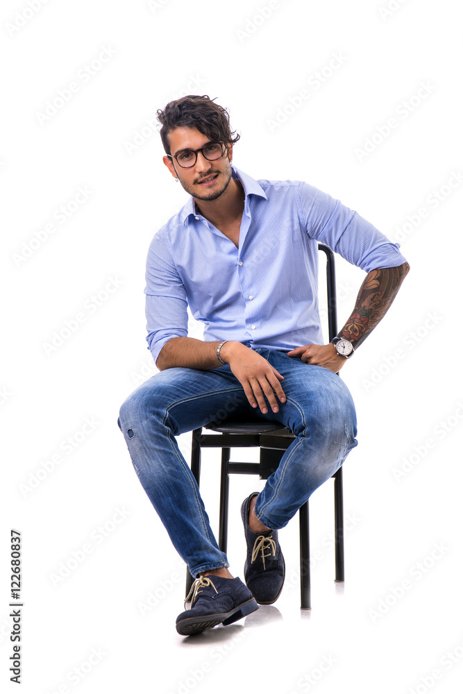 Portrait of brunette young man in light blue shirt and jeans, sitting on chair in studio shot isolated against white background. Full length photo Portrait of brunette young man in light blue shirt and jeans, sitting on chair in studio shot isolated against white background. Full length photo