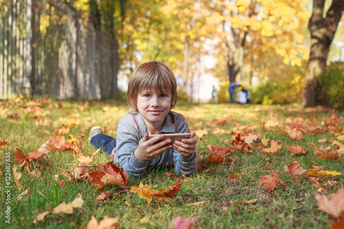 Adorable kid lying on grass in autumn park and using smartphone 