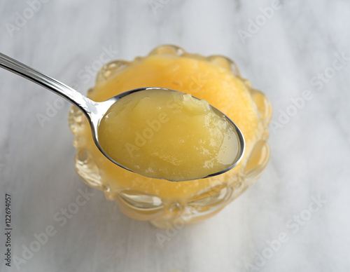 Foto Spoon full of applesauce with a glass bowl underneath atop a marble table