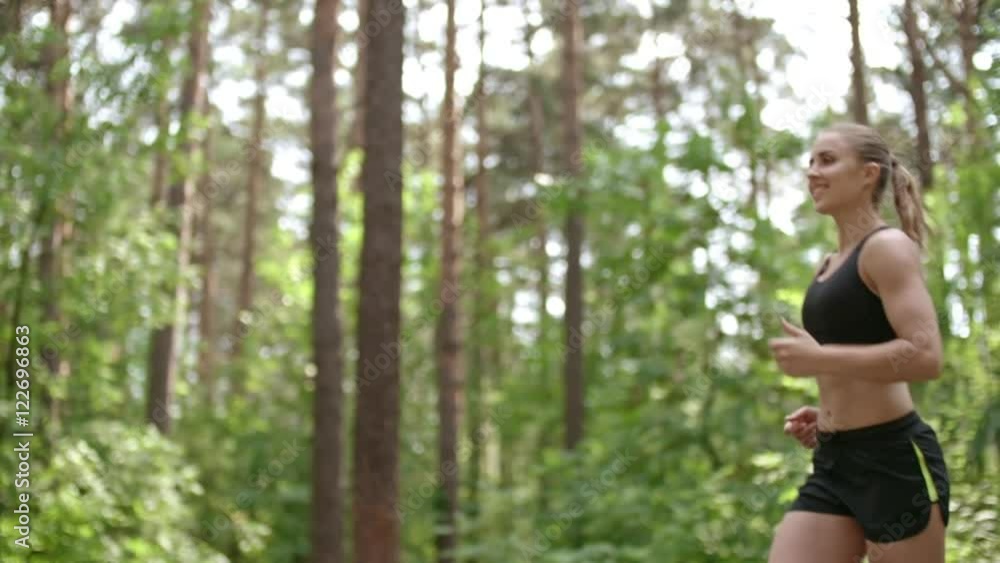 Pretty female athlete smiling while running in the forest in slow motion