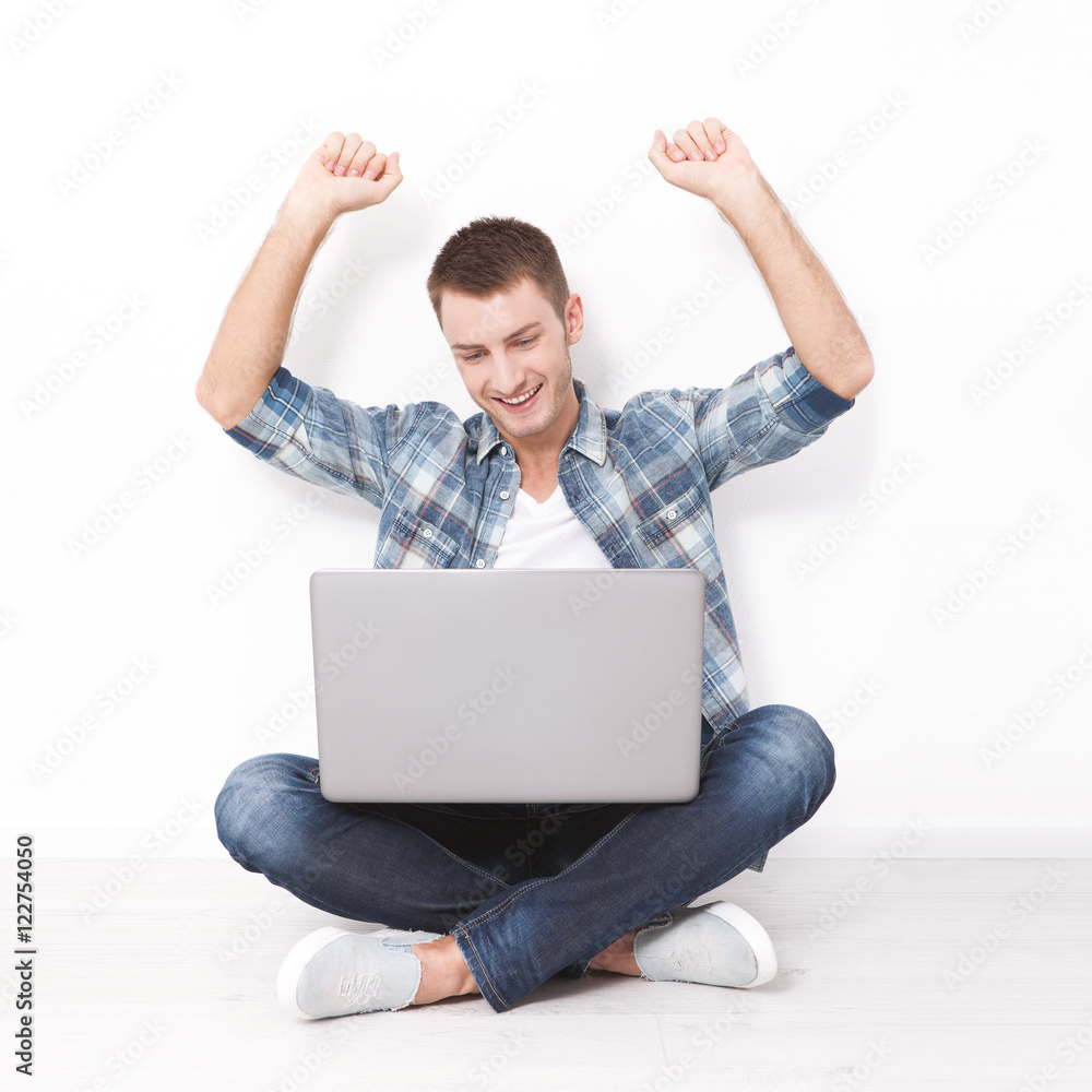 Handsome young man sitting on the floor near the wall, celebrating success with arms raised while looking at his laptop screen Handsome young man sitting on the floor near the wall, celebrating success with arms raised while looking at his laptop screen