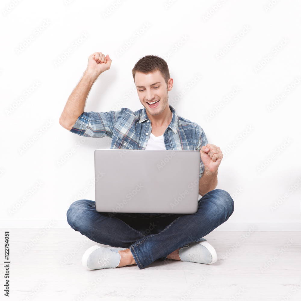 Handsome young man sitting on the floor near the wall, celebrating success with arms raised while looking at his laptop screen Handsome young man sitting on the floor near the wall, celebrating success with arms raised while looking at his laptop screen