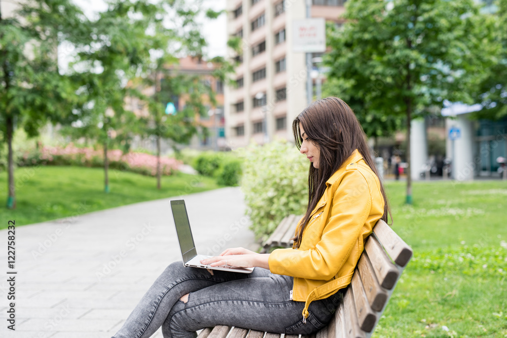 Young beautiful caucasian long straight hair woman sitting on a bench outdoor in a city park with notebook on her knee - business, technology, communication concept Young beautiful caucasian long straight hair woman sitting on a bench outdoor in a city park with notebook on her knee - business, technology, communication concept