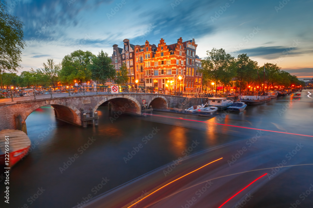 Amsterdam canal, bridge and typical houses, boats and luminous track from the boat during morning twilight blue hour, Holland, Netherlands.