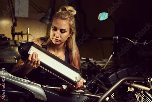 Blond woman mechanic holding a muffler in a motorcycle workshop