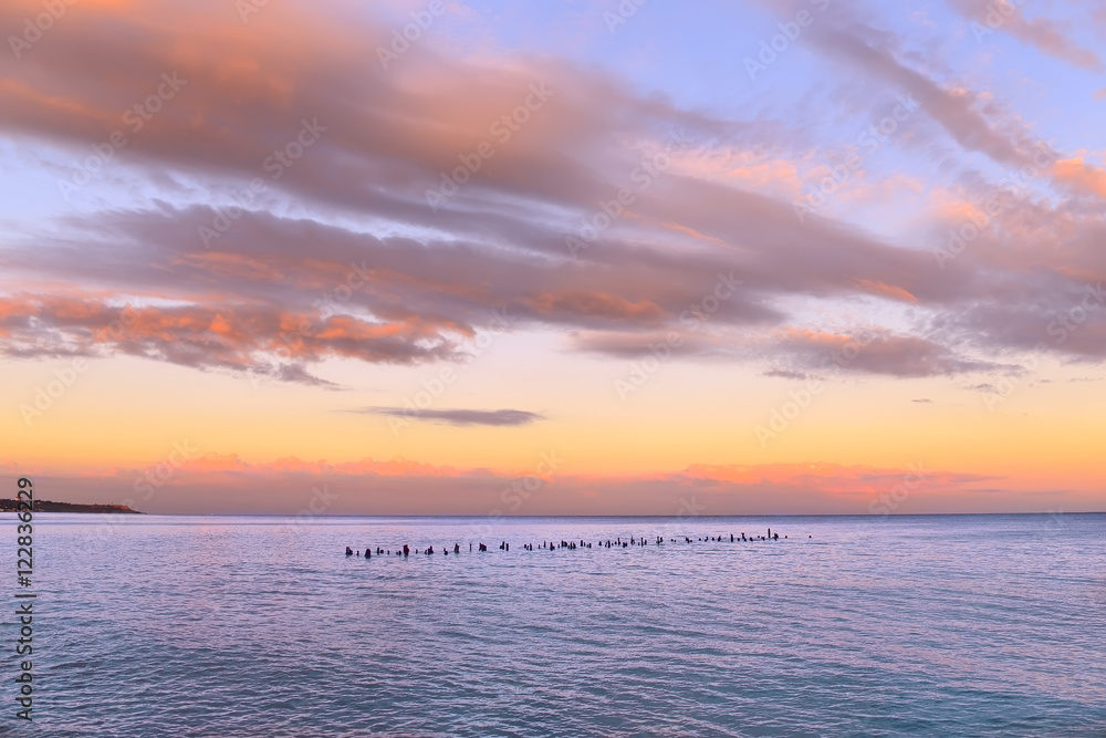 bright beautiful sunset over the sea. Red long cloud in the blue sky. In the distance is the Cape lit by the sun, in the water columns from the old wooden pier.
