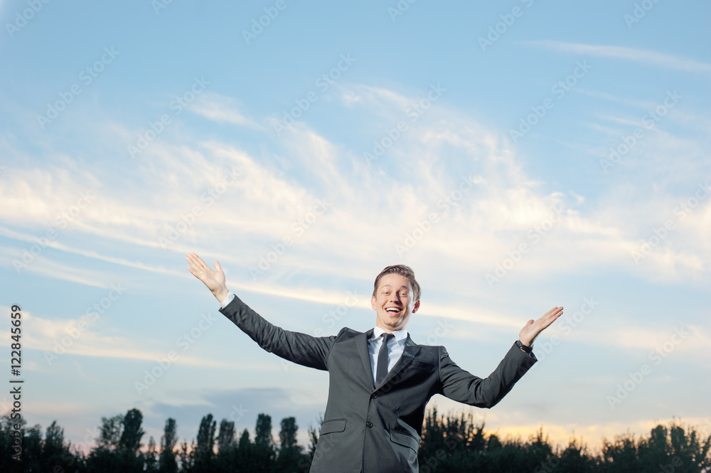 Business winner. Happy young man in formalwear keeping arms raised and expressing positivity while standing outdoors with sky in the background Business winner. Happy young man in formalwear keeping arms raised and expressing positivity while standing outdoors with sky in the background