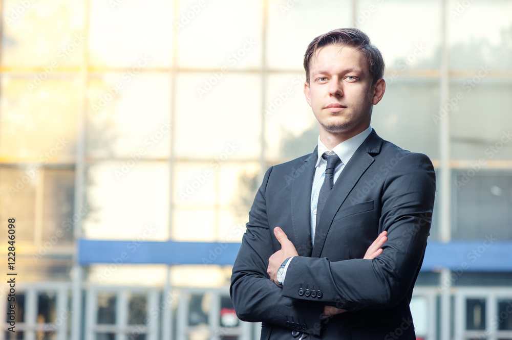 Confidence and charisma. Cheerful young businessman in full suit keeping arms crossed and looking at camera against office building Confidence and charisma. Cheerful young businessman in full suit keeping arms crossed and looking at camera against office building