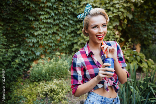 Cheerful pinup girl walking in park and drinking soda