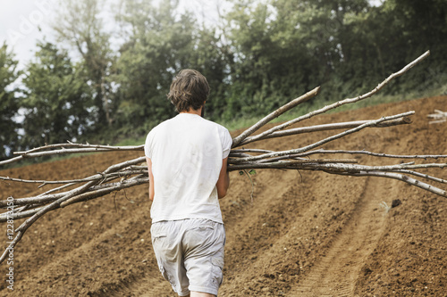 A man carrying bundles of pea sticks across tilled soil in a vegetable plot. 