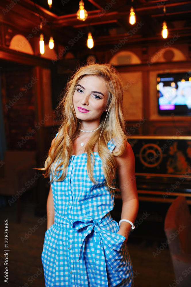 cute young blond woman in a blue sundress on a background of a bar and a variety of light bulbs