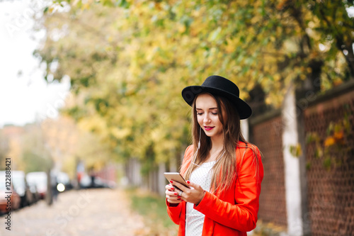young stylish pretty woman with black hat in red jacket posing in the city streets. Girl holding a phone and writing a text message