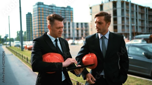 4k. Ultra hd. Two Young successful businessman customers builders in suits walk near newly constructed buildung with orange helmet and Digital Pad. Discussion about investment. Crane and beams BG photo