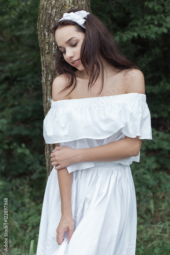 beautiful sweet girl with dark hair in a white sundress standing near a tree in the forest on hot summer day