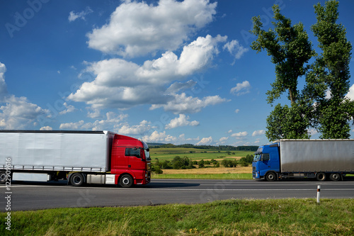 Obraz na płótnie Red truck coming together with blue truck on asphalt road with tall poplar trees in the countryside under a blue sky with white dramatic clouds. w salonie