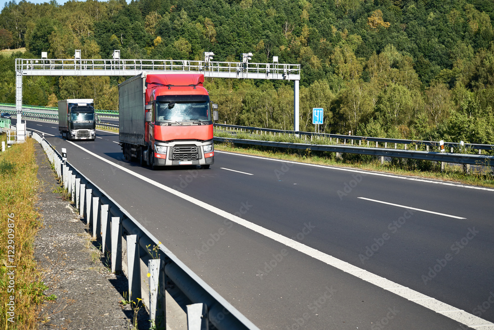 Red truck and black one passing under electronic toll collection on the asphalt highway on the forest background