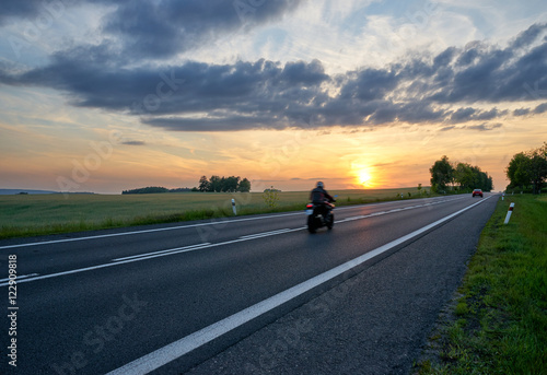 Motion blur motorcycle riding on asphalt road towards the horizon in rural landscape at sunset. Red car in the distance.