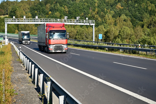 Red truck and black one passing under electronic toll collection on the asphalt highway on the forest background