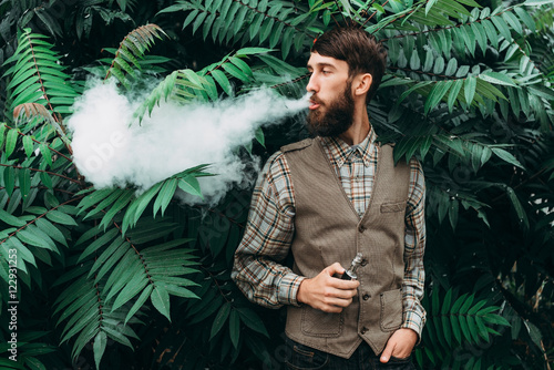 Young man with beard vaping an electronic cigarette outdoor. hipster smoke vaporizer. photo