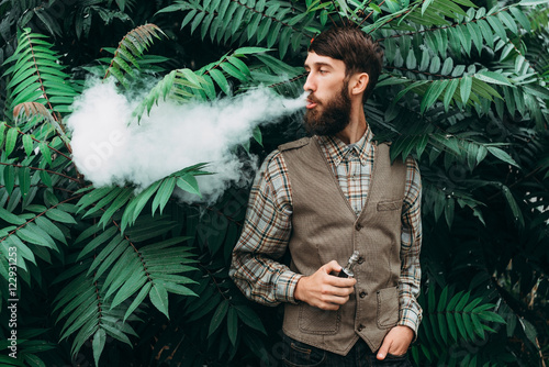 Photos Young man with beard vaping an electronic cigarette outdoor