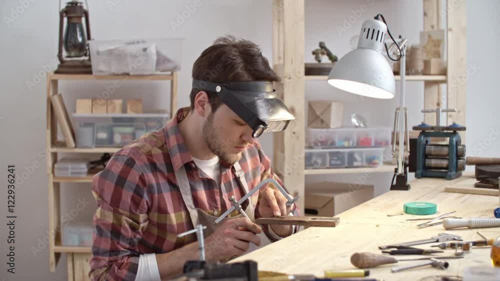 Jeweler in professional eyewear using piercing saw to cut metal on piece of wood while working in workshop Jeweler in professional eyewear using piercing saw to cut metal on piece of wood while working in workshop
