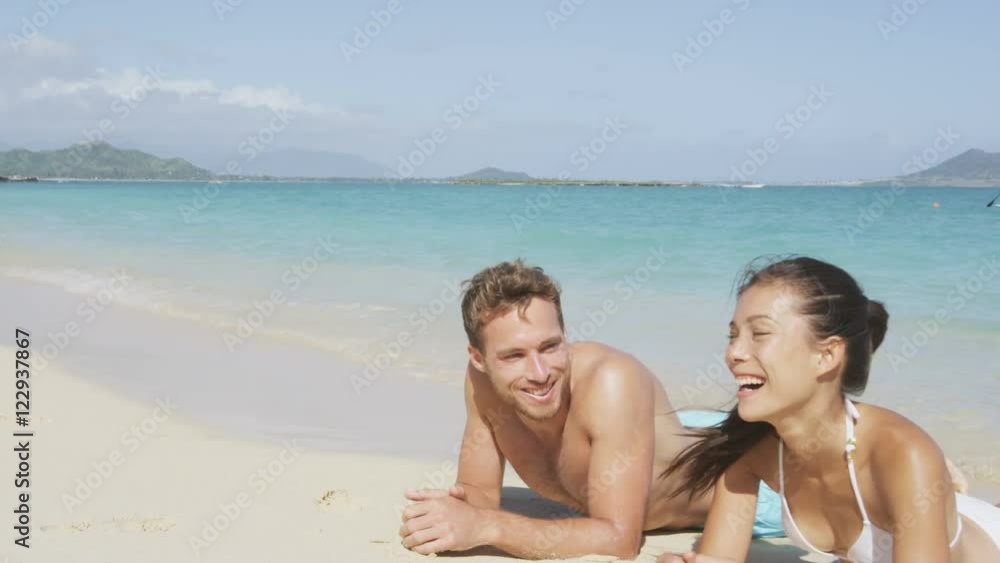 Summer beach couple on honeymoon relaxing happy laughing and smiling having fun lying down in sand. Woman and man enjoying sun tanning and summer sun on pristine paradise beach. Summer beach couple on honeymoon relaxing happy laughing and smiling having fun lying down in sand. Woman and man enjoying sun tanning and summer sun on pristine paradise beach.