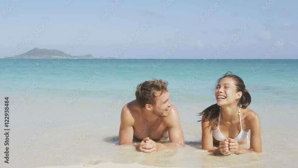 Beach couple laughing having fun with water splashing lying in sand in water edge. Woman and man on honeymoon relaxing having a good time on pristine white paradise beach. Asian woman, Caucasian man. Beach couple laughing having fun with water splashing lying in sand in water edge. Woman and man on honeymoon relaxing having a good time on pristine white paradise beach. Asian woman, Caucasian man.
