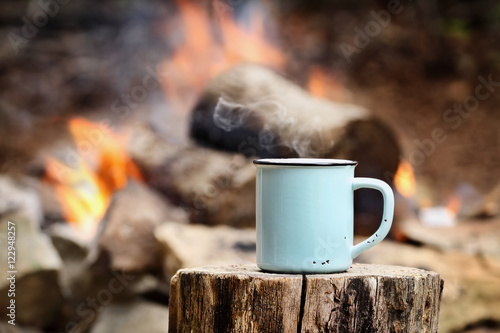 Blue enamel cup of hot steaming coffee sitting on an old log by an outdoor campfire. Extreme shallow depth of field with selective focus on mug.