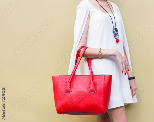 Fashionable beautiful big red handbag on the arm of the girl in a fashionable white dress, posing near the wall on a warm summer night. Warm color. Close up