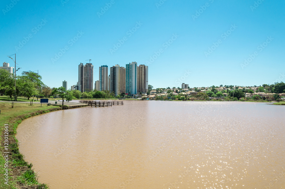 Obraz premium Lakeshore on the left with grass and some trees. Water lake and background city buildings on a sunny day.