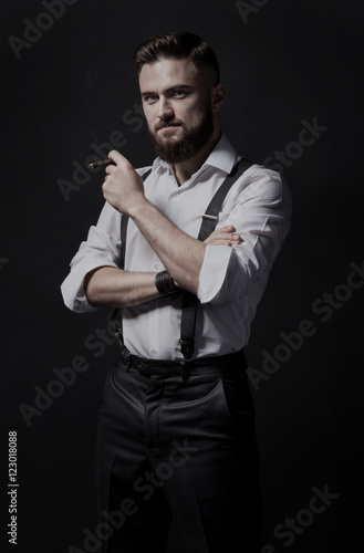 Young, stylish and attractive bearded man smoking a cigar in a dark room. He is wearing in a white shirt and suspenders. He's a smart, educated hooligan and macho