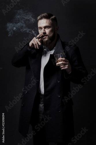 Attractive bearded man in a black coat smoking a cigar. He holds a glass with some alcohol drink. He is stylish, strong and masculine, a cloud of cigar smoke rises above it.