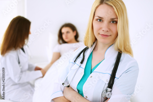 Young beautiful female doctor smiling  on the background with patient and his physician in hospital. High level and quality medical service concept.