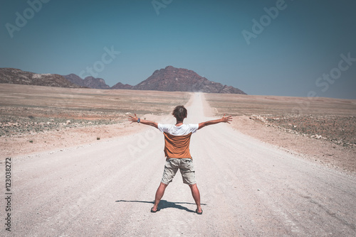 Woman with outstretched arms standing on gravel road crossing the Namib desert, in the Namib Naukluft National Park, main travel destination in Namibia, Africa. Rear view, toned image.